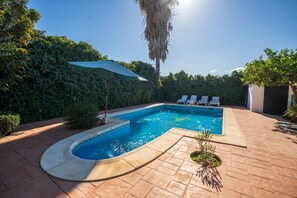 Ein rechteckiger Pool mit geschwungener Kante, umgeben von einer Ziegelterrasse. Liegestühle und ein grüner Sonnenschirm sind vorhanden.