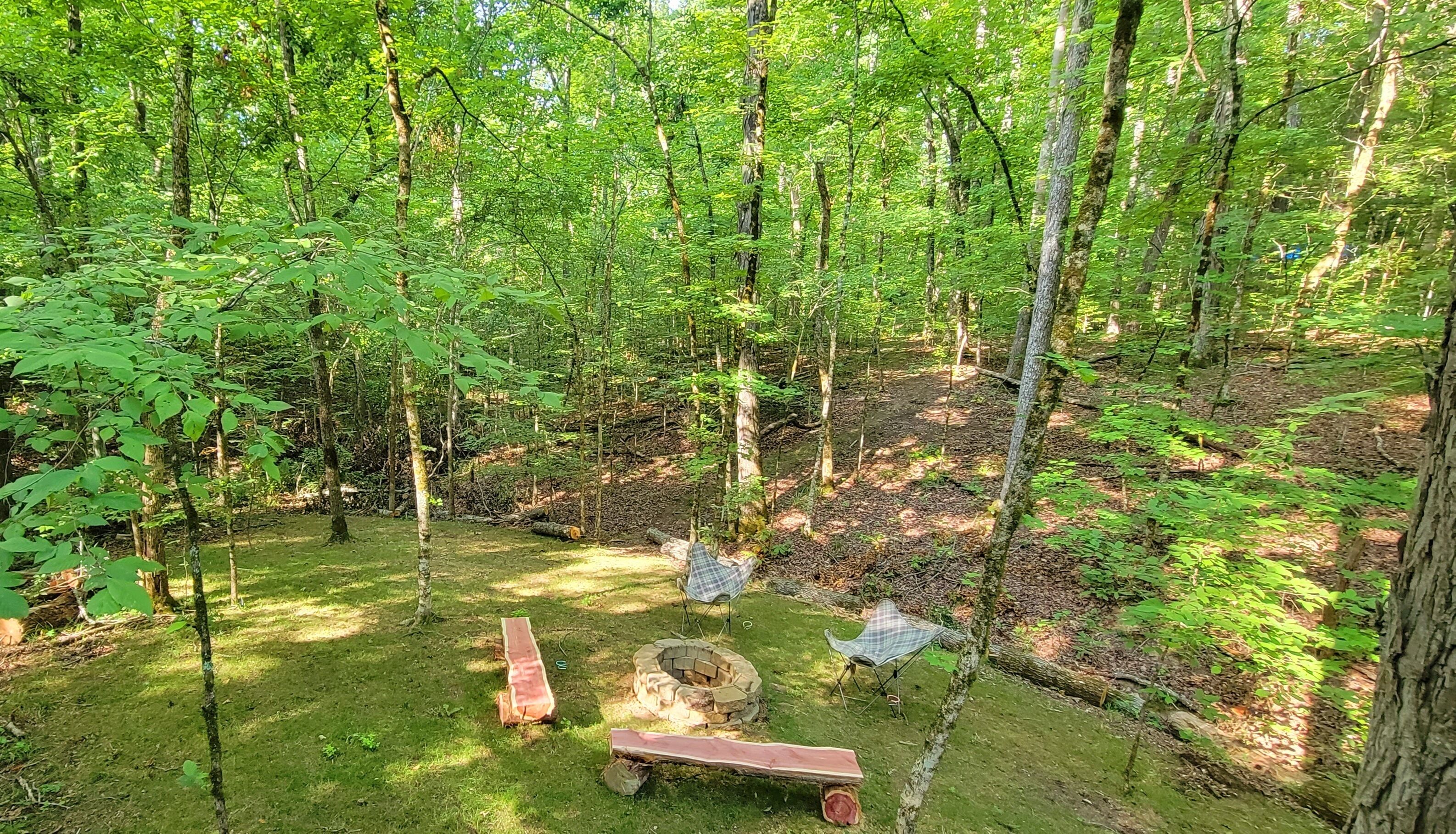 A forested campsite with a fire pit, picnic table, and chairs.
