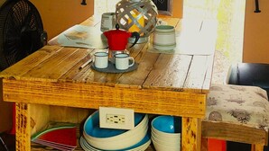 Een rustieke houten tafel met een koffiepot, mokken en borden. Er zijn ramen met gordijnen en een ventilator op de achtergrond.