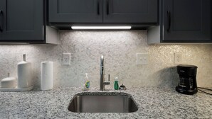 A kitchen with a stainless steel sink, dark cabinets, a herringbone-patterned backsplash, and a coffee maker on the counter. - Soho Loft 5 (Cartersville)