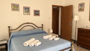 Een keurig opgemaakt bed met blauw bedlinnen en opgerolde handdoeken, een houten garderobe, een nachtkastje met lamp en drie ingelijste foto's aan de muur.