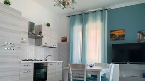 Een moderne keuken met witte kasten, een gasfornuis en een inbouwoven. Er is een eethoek met een witte tafel en stoelen, en een flatscreen televisie aan de muur bevestigd.