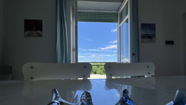 Een kamer met een tafel en stoelen, een raam met gordijnen, en uitzicht op de lucht en bomen.
