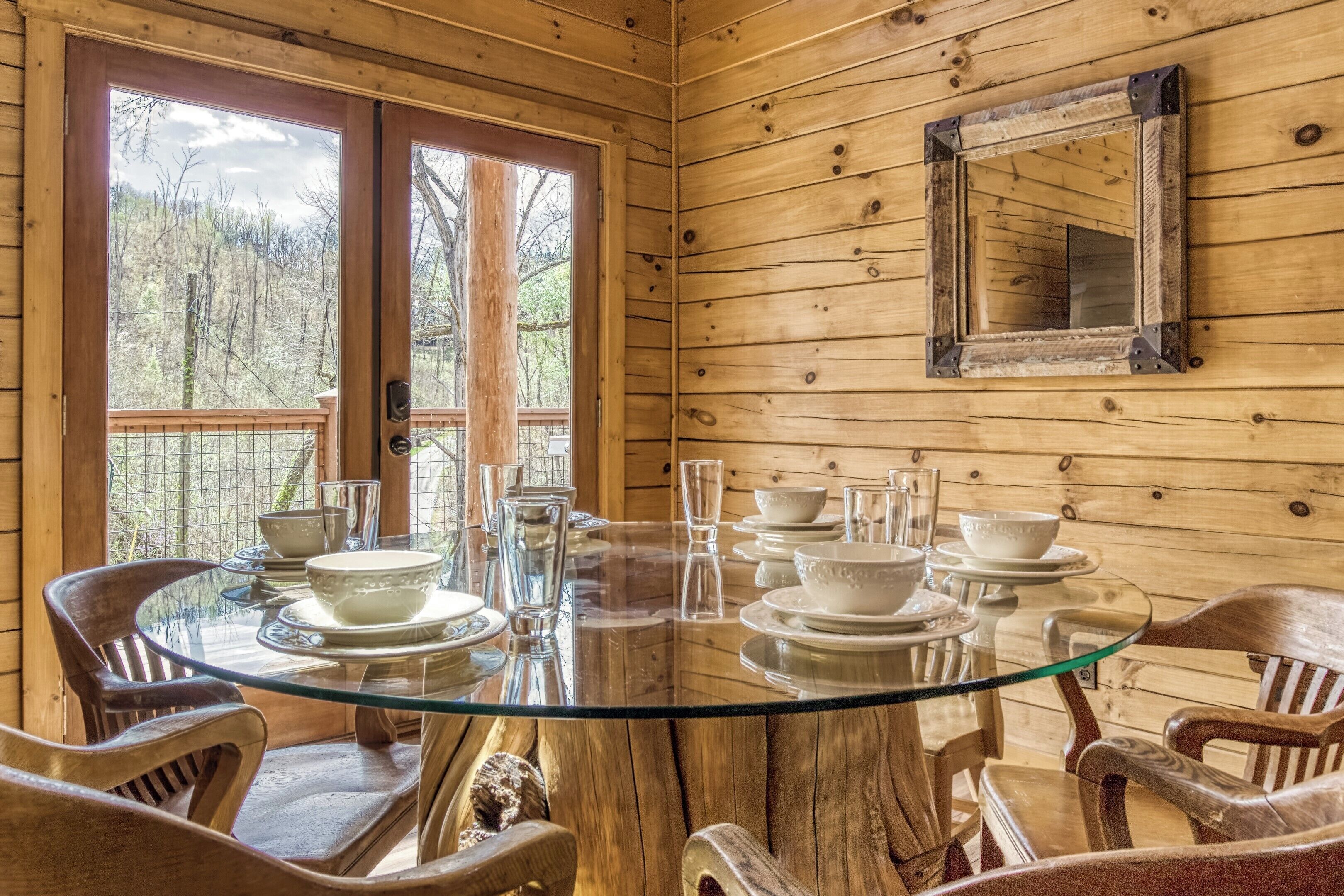 Interior de una cabaña de madera con una mesa de comedor de vidrio preparada para cuatro personas, rodeada de sillas de madera.