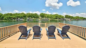 A deck with four Adirondack chairs facing a lake, wooden railings, and a clear view of the water and distant buildings.