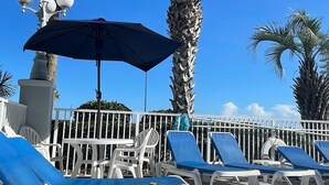 Un área junto a la piscina con sillas reclinables azules, una sombrilla grande y palmeras.