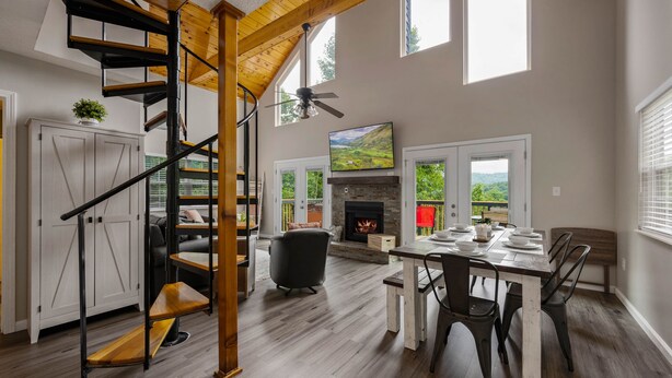 A spacious living area with a wooden staircase, a fireplace, a dining table, and a large window offering a view of the outdoors. - Stylish Mountain Retreat w/ Hot Tub, Games and Stunning Sunrise Mountain Views! (Sevierville)