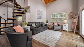 A spacious living room with a black leather sofa, a wooden spiral staircase, and a large window with blinds. - Stylish Mountain Retreat w/ Hot Tub, Games and Stunning Sunrise Mountain Views! (Sevierville)