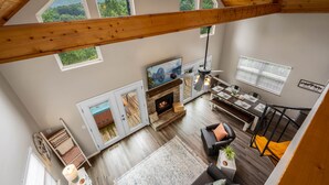 A spacious loft with a high ceiling, wooden beams, a fireplace, a dining area, and large windows offering a view of the outdoors. - Stylish Mountain Retreat w/ Hot Tub, Games and Stunning Sunrise Mountain Views! (Sevierville)