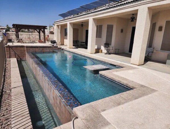 A swimming pool with a tiled edge, a pergola, and a covered patio area with chairs.
