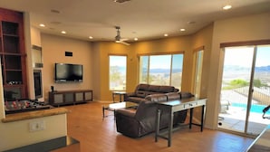 A spacious living room with a sofa, coffee table, and a foosball table. There is a TV mounted on the wall and large windows offering a view of the outdoors. - Custom 4 bdrm with Granny Suite and Infinity Pool (Bullhead City)