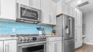 A kitchen with a stainless steel microwave, oven, and refrigerator, light blue backsplash, and white cabinets.