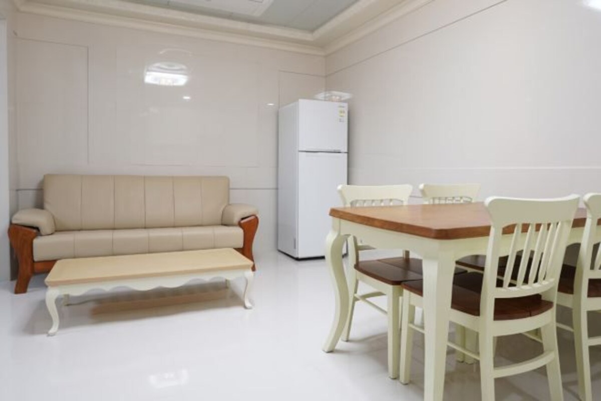 A room with a beige sofa, a wooden coffee table, a white dining table with four chairs, and a white refrigerator.