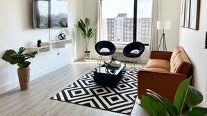 A modern living room with a large window, a flat-screen TV, a sofa, and a coffee table. - Stylish King 1BR Near MetLife & NYC (Elizabeth)