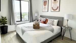 A modern bedroom with a large bed, two bedside lamps, a view of the city through a window, and two framed artworks on the wall. - Stylish King 1BR Near MetLife & NYC (Elizabeth)
