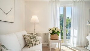 Um quarto em tons de branco, com um sofá branco, uma mesinha com uma planta, um radiador e uma janela com cortinas.