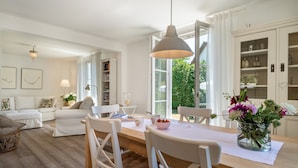 Uma sala de estar moderna com uma mesa de jantar de madeira, mobiliário branco e uma janela grande com vista para vegetação.