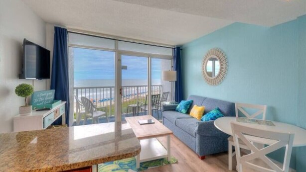 A modern living room with a blue wall, a grey sofa with colorful cushions, a white dining table, and a large window offering a view of the sea. - Seawatch Resort - King Bed (Myrtle Beach)