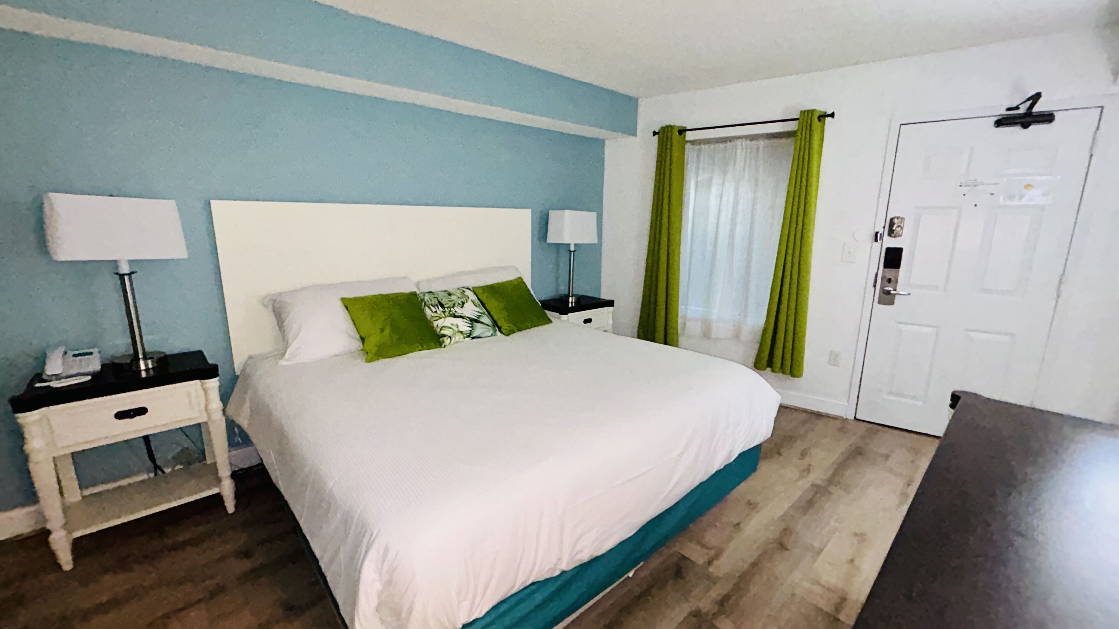 Une chambre d’hôtel comprenant un lit, deux tables de chevet, une commode, une lampe, une porte et une fenêtre avec des rideaux.