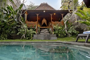 A traditional wooden structure with a tiled roof, surrounded by lush greenery and a clear swimming pool. - Bubu Mesari Ubud Villa (Ubud)