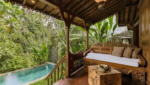 Una terraza de madera con piscina, rodeada de exuberante vegetación y un banco de madera con cojines.