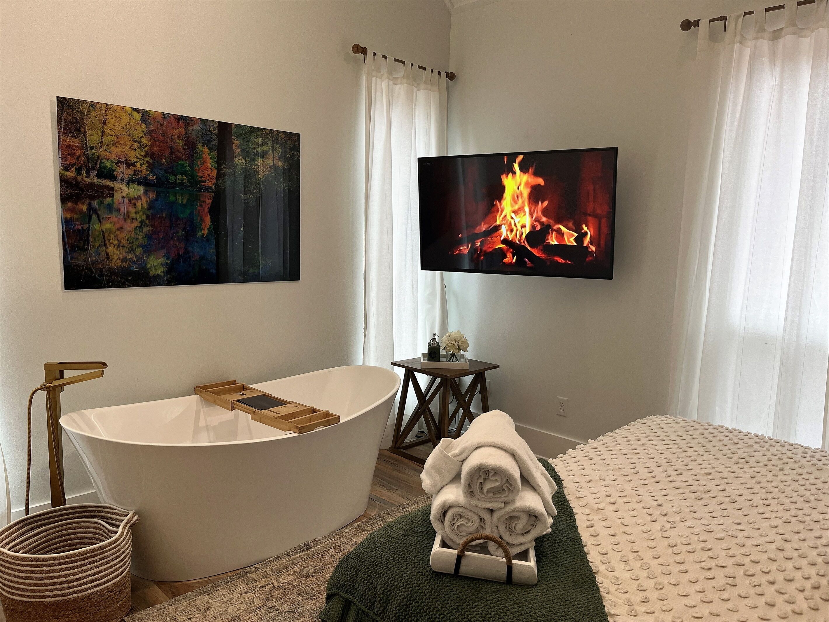 Un baño moderno con una bañera independiente, un televisor de pantalla plana mostrando un fuego, y un cuadro con una escena de bosque.