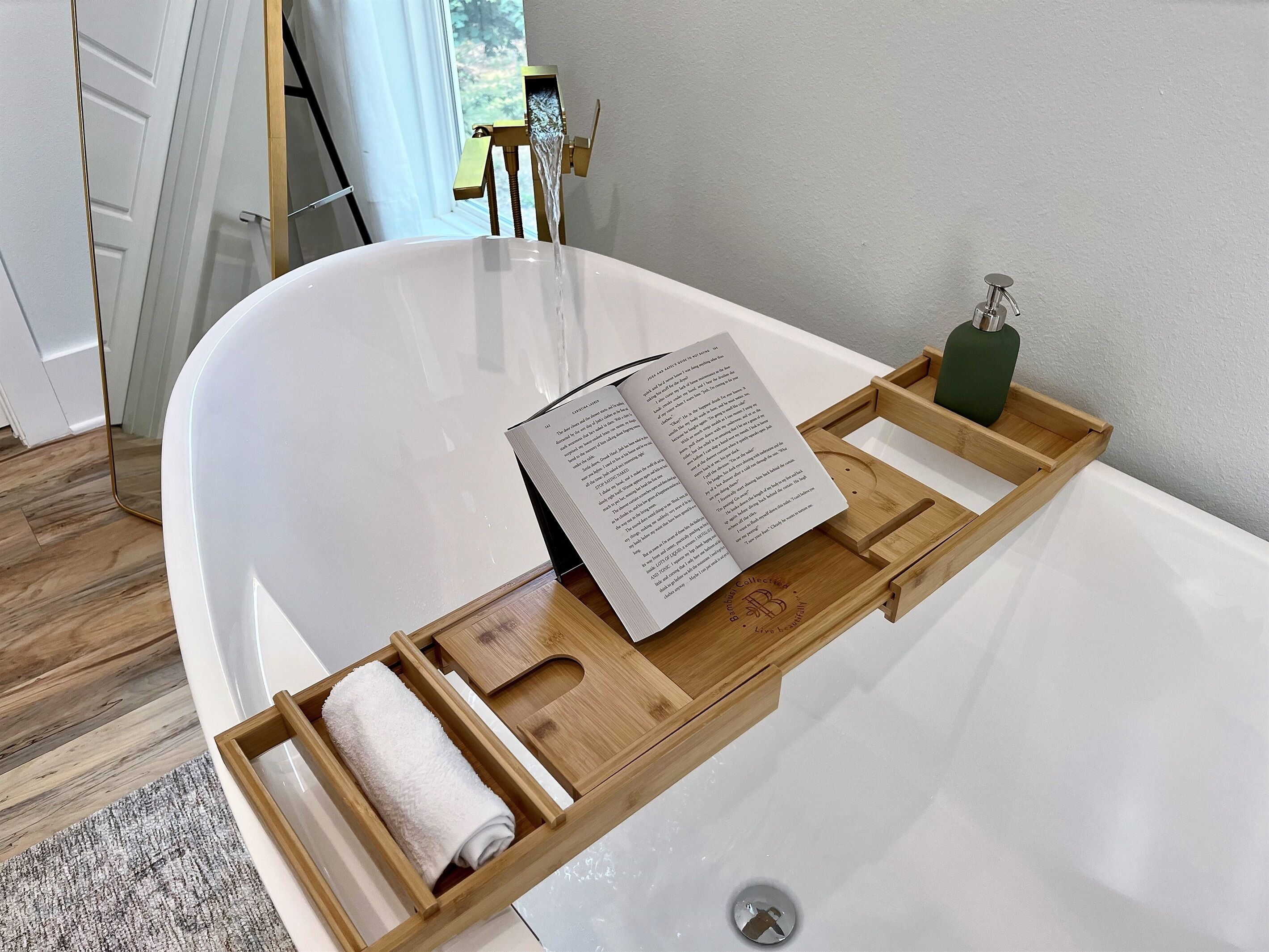 Un baño moderno con una bañera blanca, una bandeja de madera con un libro y una toalla, y un dispensador de jabón verde.