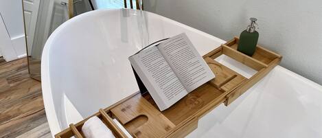 Un baño moderno con una bañera blanca, una bandeja de madera con un libro y una toalla, y un dispensador de jabón verde.