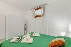 Ein Hotelzimmer mit grüner Bettdecke, einem weißen Kleiderschrank, einem Fenster mit Vorhängen, einer Nachttischlampe und einem Handtuch auf dem Bett.