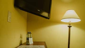 Habitación de hotel con televisión de pantalla plana montada en la pared, una mesa con lámpara y agua embotellada.