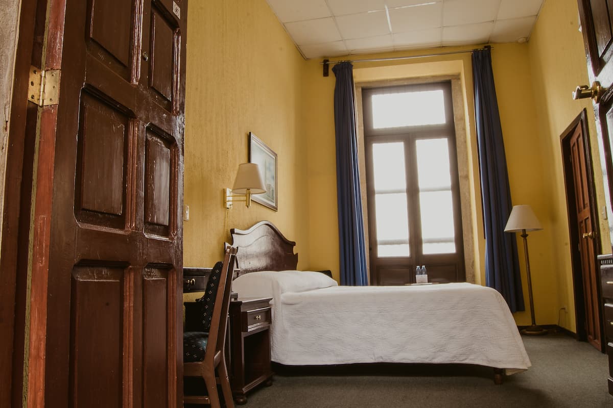 Habitación de hotel con cama, escritorio, silla, lámpara, ventana con cortinas y puerta.