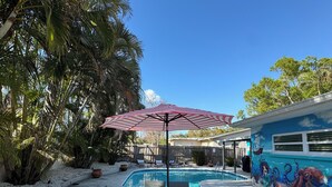 A swimming pool with a red umbrella, surrounded by palm trees and a mural of an octopus on the building.