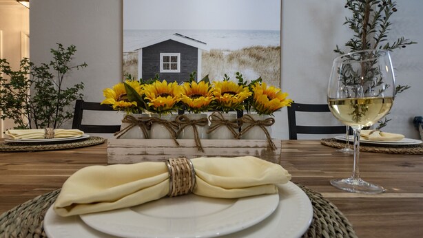 A dining table set with white plates, a folded napkin, and a glass of white wine. Sunflowers are arranged in a vase on the table.