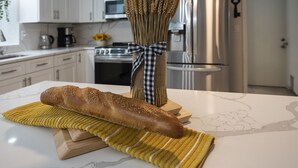A kitchen with white cabinets, a stainless steel refrigerator, and a wooden cutting board with a baguette and a vase of wheat stalks.