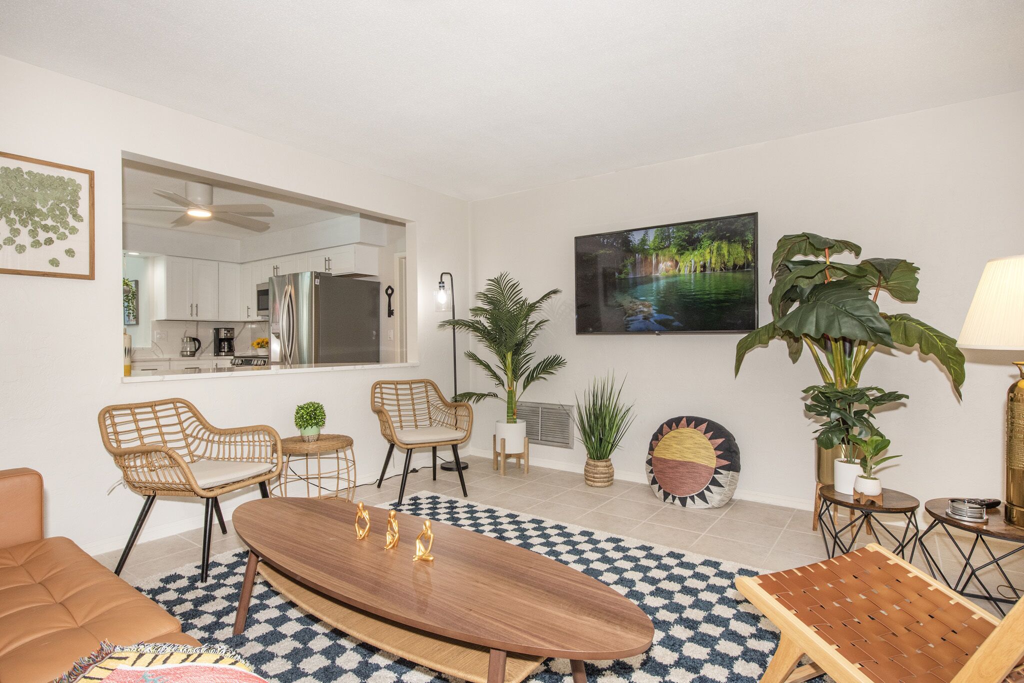 A modern living room with a checkered rug, a wooden coffee table, and a sofa. There are two wicker chairs, a dining table, and a large potted plant. A kitchen is visible in the background.
