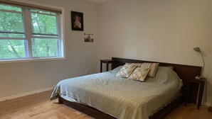 Un dormitorio con piso de madera, una cama con ropa de cama estampada, dos mesitas de noche y una ventana con vistas a árboles.