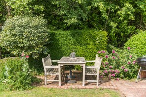 A garden patio with a table and chairs, surrounded by lush greenery and flowering plants. - Charming countryside cottage with large garden (Hammill)