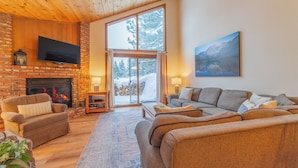 Una sala de estar acogedora con chimenea, un televisor montado en una pared de ladrillo y un ventanal con vistas a árboles cubiertos de nieve.