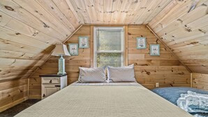 A cozy attic bedroom with wooden walls, a bed with a beige blanket, a nightstand with a lamp, and framed pictures on the wall.