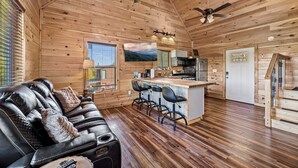 A cozy cabin interior with a wooden ceiling, a black leather sofa, a kitchen area with bar stools, and a television mounted on the wall.