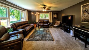 A living room with a sectional sofa, a piano, a television, and a ceiling fan. - Dog-Friendly Boyne City Home | Game Room, Near Lake Charlevoix + Downtown (Boyne City)