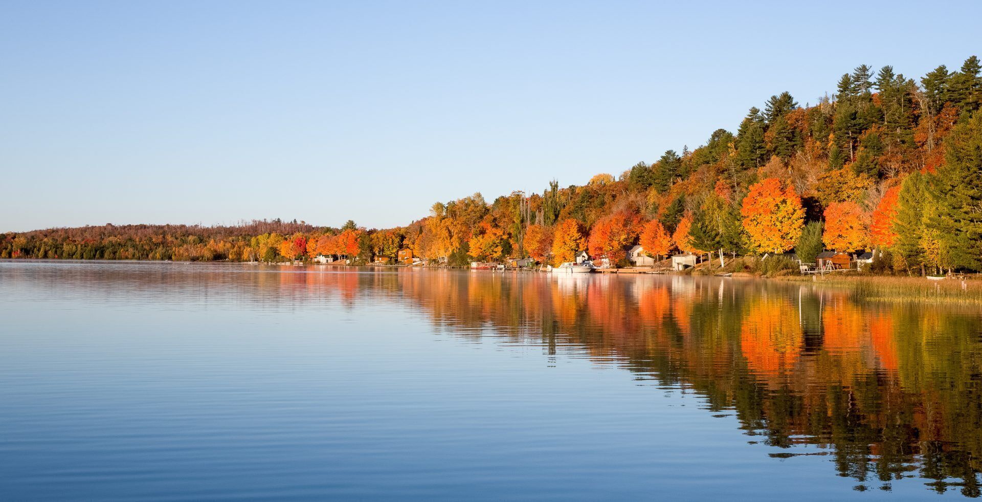 Een kalm meer met herfstkleurige bomen, hun reflectie in het water, omringd door een bos en een rij huizen.