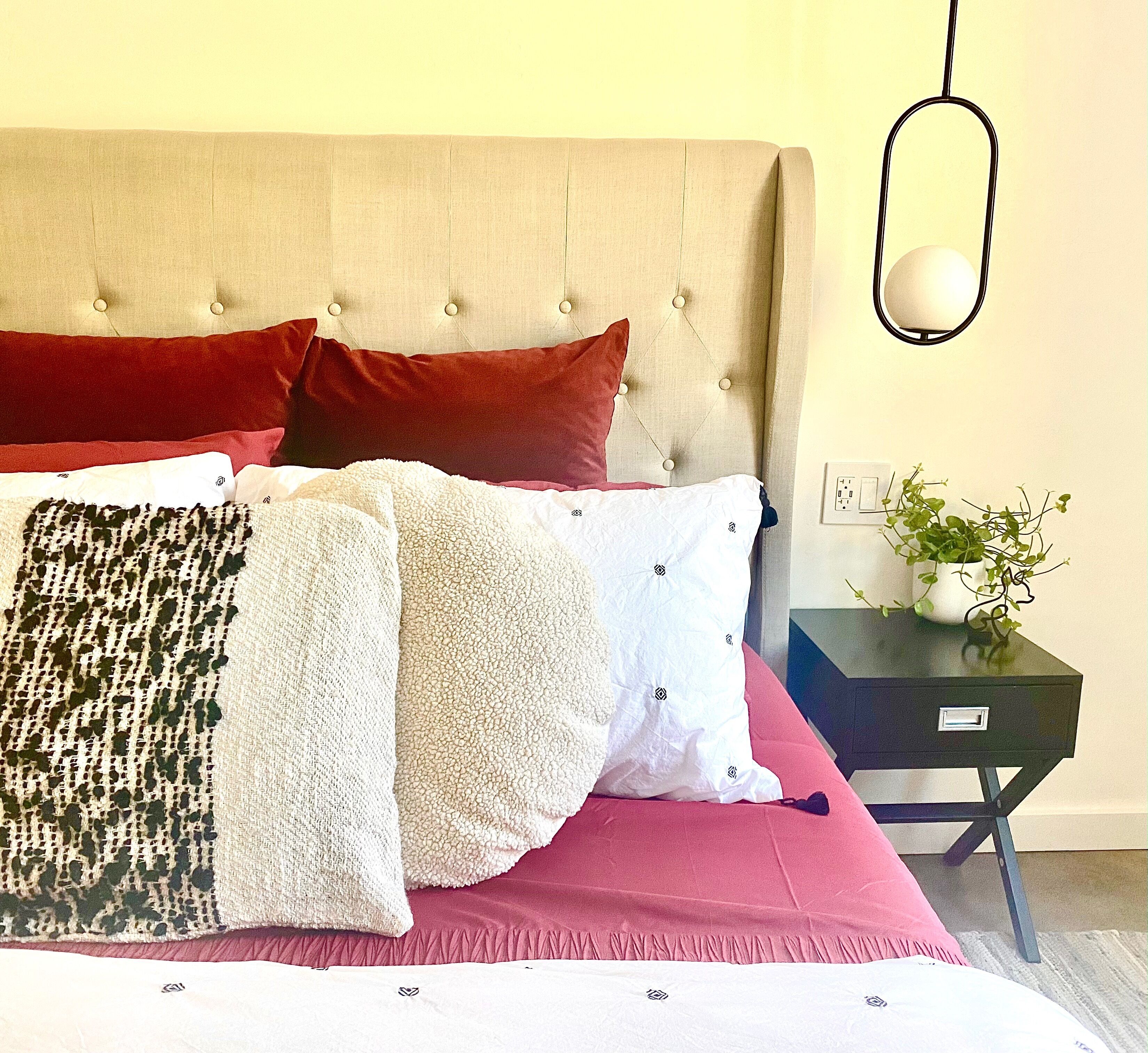 Una cama bien hecha con varias almohadas y una colcha, una mesita de noche con una planta, y una lámpara fijada a la pared.