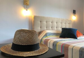A bedroom with a tufted headboard, a striped bedspread, a woven hat, and two wall-mounted lights.