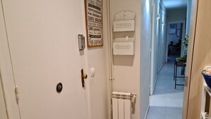 Un couloir avec une porte blanche, un radiateur et un panneau fixé au mur indiquant différentes catégories, comme « LETTRES » et « MAGAZINES ».