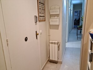 Un couloir avec une porte blanche, un radiateur et un panneau fixé au mur indiquant différentes catégories, comme « LETTRES » et « MAGAZINES ».
