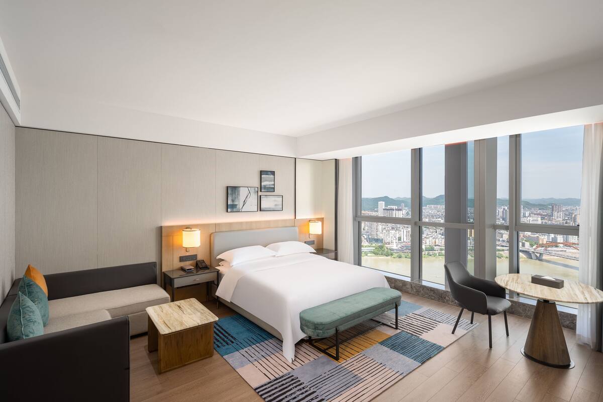 A modern hotel room with a large bed, a sofa, a small table, and a view of the city through large windows.