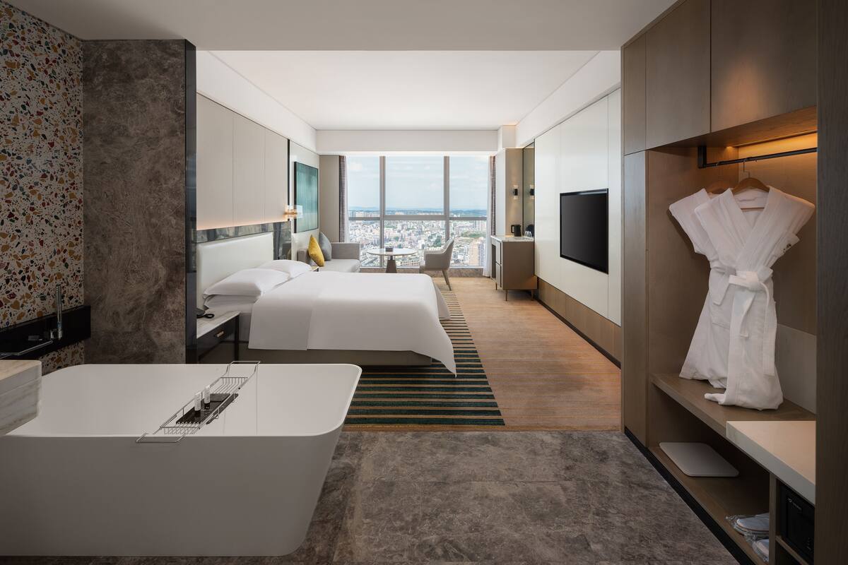 A modern hotel room with a large bed, a separate seating area, a TV, and a view of the city.