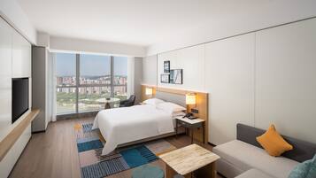 A modern hotel room with a large bed, a sofa, a small table, a TV, and a city view.