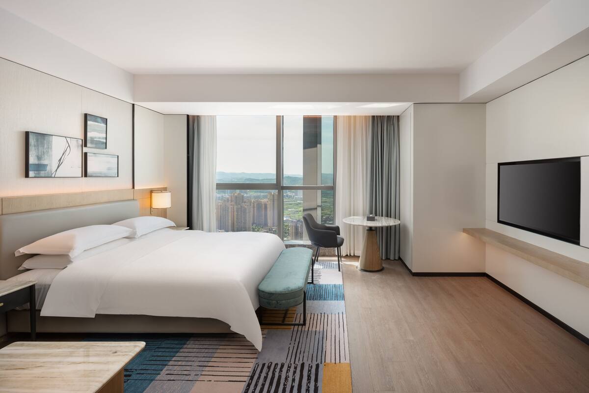 A modern hotel room with a large bed, a seating area, a TV, and a view of the city.